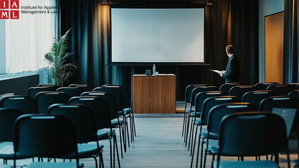 A trainer stands at the podium in a seminar room, featuring rows of chairs facing a presentation screen.